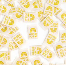 Load image into Gallery viewer, Woven Labels by Sarah Hearts white background with gold rainbow and Hecho por Mi or Made by Me in Spanish in gold for clothing garments purses tote bags quilts cotton washable