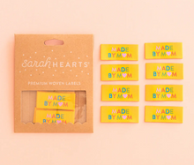 Load image into Gallery viewer, Woven end fold labels by Sarah Hearts bright yellow background with Made by Mom in rainbow colors of turquoise green orange pink with pink heart instead of an O in Mom used for clothing garments quilts costumes bags totes purses crafts