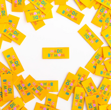 Load image into Gallery viewer, Woven end fold labels by Sarah Hearts bright yellow background with Made by Mom in rainbow colors of turquoise green orange pink with pink heart instead of an O in Mom used for clothing garments quilts costumes bags totes purses crafts