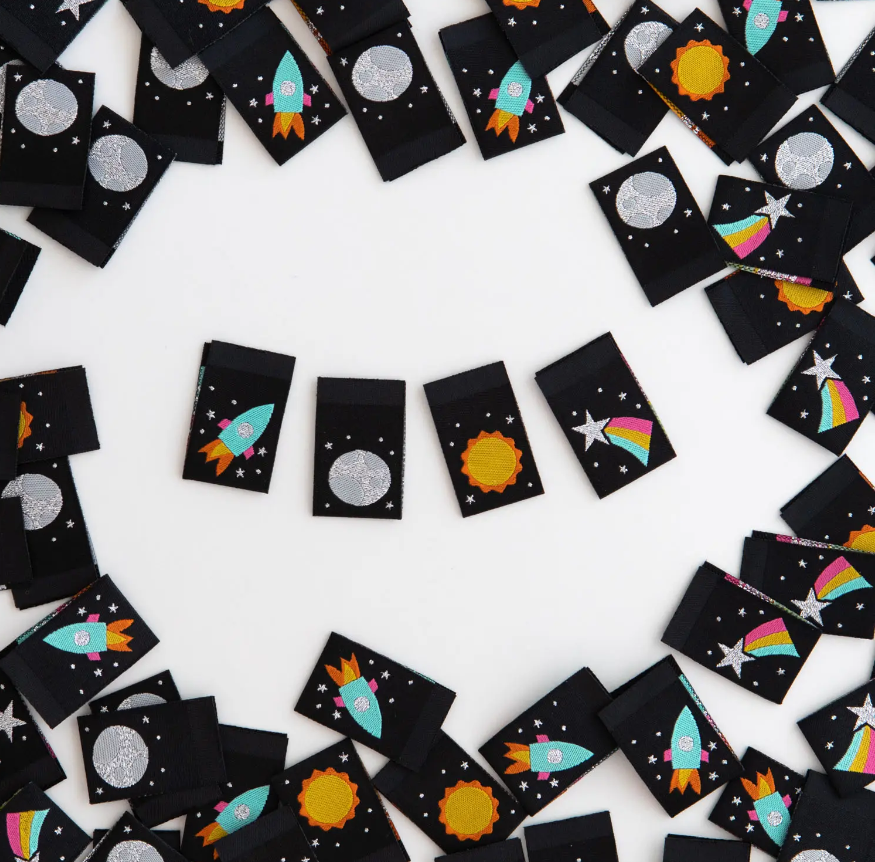 Woven space themed centerfold cotton organic label by Sarah Hearts  4 designs on black background with an aqua rocket ship orange flames and silver stars a silver full moon and sprinkled stars yellow and orange sun and silver shooting star with rainbow tail use for clothing garments bags gifts quilts zippies washable organic cotton